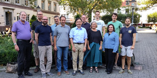 12 Personen stehen für die Fotoaufnahme auf einem Parkplatz. Im Hintergrund sind alte Gebäude zu sehen.