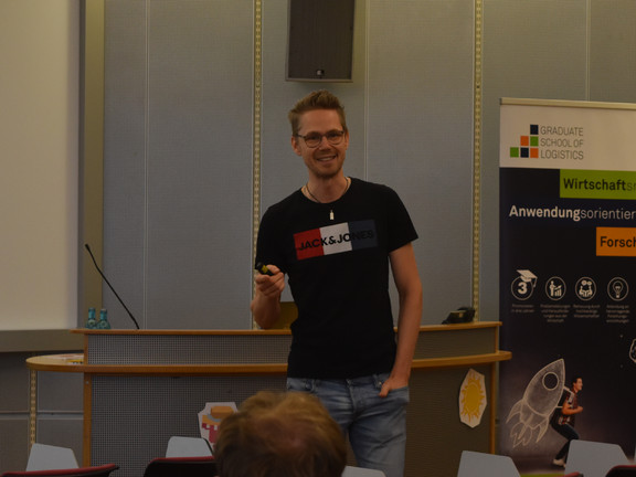 Ein Mann steht in einem Hörsaal vor einer Präsentation und spricht zu den Zuhörenden. Im Hintergrund ist in einer Ecke ein Roll-Up zu sehen.
