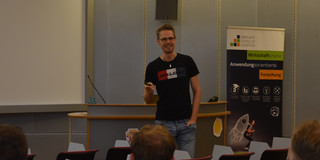 Ein Mann steht in einem Hörsaal vor einer Präsentation und spricht zu den Zuhörenden. Im Hintergrund ist in einer Ecke ein Roll-Up zu sehen.