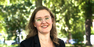 Eine Frau sitzt auf einer Astgabel und schaut in die Kamera. Im Hintergrund stehen Bäume.